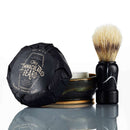 A shaving brush stands next to a ceramic dish with a lid labeled The Immaculate Beard Immaculate Dense Shave Soap For Smooth Men Wet Shaving, making it ideal for a traditional wet shave. The set is displayed on a reflective white surface.