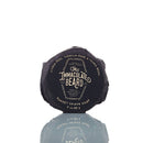 A round, dark-wrapped bar of The Immaculate Beard Dark Shave Soap Puck, this natural shave soap features text describing its scent—citrus peel, vanilla musk, and tonka bean—against a white background.