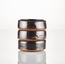 A small, dark brown ceramic Shave Bowl by The Immaculate Beard, featuring a glossy finish and three distinct ridges, sits on a white, reflective surface—perfect for whipping up a rich, moisturizing lather from your favorite shave soap puck.