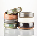 Eight Shave Bowls by The Immaculate Beard in earthy ceramic tones, stacked in two uneven piles on a reflective surface, each showcasing unique texture and glaze variations—perfect for creating a rich, moisturizing lather with your favorite shave soap puck.