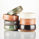 A stack of The Immaculate Beard Shave Bowls in earthy tones—green, brown, black, and white—is displayed on a plain white background with a subtle reflection beneath them. These handmade ceramic bowls are ideal for creating a rich, moisturizing lather with your favorite shave soap puck.