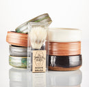 A boxed The Immaculate Beard shaving brush is displayed in front of a neatly stacked set of eight colorful Shave Bowls by The Immaculate Beard, arranged in two columns on a white background. Each handmade Shave Bowl is perfect for building up a rich, moisturizing lather with your favorite shave soap puck.