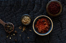 A bowl of World Spice Shawarma Spice, a small bowl of cumin seeds, a copper spoon filled with black peppercorns, and a bowl of vibrant red spice from World Spice are artfully arranged on a rustic black charred wooden surface, evoking the richly flavored ingredients often found in Middle Eastern spit-roasted dishes.