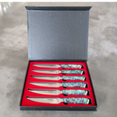The BBQ Boards® BBQ Blades™ Steak Knife Set features six premium steak knives with ornate patterns and colorful handles, elegantly displayed in an open black box lined with red felt on a gray surface.