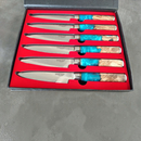 The BBQ Blades™ Steak Knife Set by BBQ Boards® features six premium steak knives with shiny blades and wood-and-blue-resin handles, elegantly arranged in a red-lined black box.