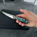 A hand holds a BBQ Blades™ Steak Knife by BBQ Boards®, featuring a serrated, patterned blade and an iridescent handle on a black surface—an elegant piece from their premium steak knife set.