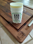 An open tube of BBQ Boards® BBQ Board Balm® Care Pair Wax and Oil, labeled Rosemary Lime, rests on a wooden cutting board engraved with the BBQ Boards® logo. The scene is set atop tiled flooring.