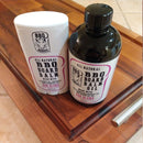 Two BBQ Boards® “BBQ Board Balm® Care Pair Wax and Oil” containers—a black oil bottle and a white wax stick, both with purple/black labels—sit on a wooden cutting board, ideal for cutting board care.