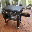 The BBQ Boards® 30" Blackstone Griddle Side Cutting Board features a closed lid and a side cutting board, shown on a wooden deck with patio furniture and plants—ideal for flat top grill prep.