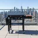 A BBQ Boards® 30" Blackstone Griddle Side Cutting Board by BBQ Boards® is set on a rooftop terrace with a city skyline backdrop, providing ample flat-top prep space for all your BBQ cutting board needs.
