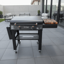 A Blackstone outdoor griddle grill with two knobs and a BBQ Boards® 30" Blackstone Griddle Side Cutting Board sits on a modern patio with gray tiles, surrounded by outdoor furniture and potted plants.