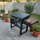 A BBQ Boards® 30" Blackstone Griddle Side Cutting Board is attached to a black grill on a stone patio, surrounded by potted flowers and a wicker chair. Wooden fencing and green shrubs provide the perfect flat top prep backdrop.