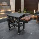 A BBQ Boards® 30" Blackstone Griddle Side Cutting Board is set up on a modern outdoor griddle with two knobs, closed lid, and lower storage shelf, on a gray stone patio near water and seating.
