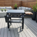 A BBQ Boards® 30" Blackstone Griddle Side Cutting Board sits next to a Blackstone outdoor griddle with a flat top prep area on a spacious wooden deck, surrounded by modern patio furniture, potted plants, and wooden privacy fencing.