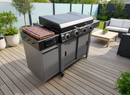 A modern outdoor gas grill with four burners and a BBQ Boards® 36" Blackstone Griddle Side Cutting Board is set on a wooden patio, surrounded by potted plants, garden furniture, a table, and cushioned seating.