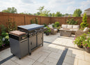 A modern gas grill on a stone patio with wicker furniture, coffee table, blooming flowers, and a wooden fence features the BBQ Boards® 36" Blackstone Griddle Side Cutting Board—perfect for meal prep amid green trees and blue sky.