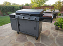 A BBQ Boards® 36" Blackstone Griddle Side Cutting Board enhances a large, gray outdoor gas grill with four knobs on a stone patio in the backyard, surrounded by green grass, potted plants, and patio furniture.