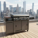 A stainless steel gas grill with four burners stands on a rooftop deck, featuring the BBQ Boards® 36" Blackstone Griddle Side Cutting Board, with city skyscrapers in the background under a clear sky.