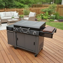 A large, modern gas grill with four burners stands on a wooden deck, featuring storage cabinets and a BBQ Boards® 36" Blackstone Griddle Side Cutting Board. An outdoor seating area and garden appear in the backyard background.