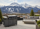 A modern patio with a BBQ Boards® 36" Blackstone Griddle Side Cutting Board, fire pit, and outdoor sofa set overlooks a town and snow-capped mountains with ski slopes beneath a partly cloudy sky.