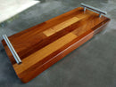 The BBQ Boards® Baby Block 18" x 8" Cutting Board features a pattern of different wood tones and sturdy metal handles, blending elegance and practicality in a compact design. The glossy finish adds style while resting on a smooth gray surface.