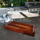 The BBQ Boards® Baby Block 18" x 8" Cutting Board sits on a gleaming outdoor countertop with a built-in sink, faucet, and barbecue grill in the background—perfect for serving snacks or prepping food.