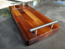 The BBQ Boards® Baby Block 18" x 8" Cutting Board features a patchwork wooden design and sleek metal handles, resting on a gray surface. Sunlight highlights its glossy finish, adding elegance and versatility to this compact board.