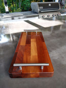 The BBQ Boards® Baby Block 18" x 8" Cutting Board with metal handles rests on a polished outdoor counter, with a gas grill and greenery in the background.
