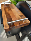 A close-up of a BBQ Boards® Baby Block 18" x 8" Cutting Board attached atop a smoker grill, featuring two metal handles and the BBQ Boards® logo in the corner.