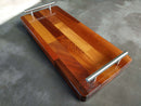 The BBQ Boards® Baby Block 18" x 8" Cutting Board features a rectangular patchwork of wood tones and two metal handles, doubling as a serving tray, shown on a gray surface.