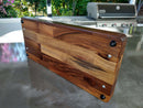 The Baby BBQ Board® 18" x 8" Cutting Board by BBQ Boards® stands upright on a sleek gray outdoor countertop. Black rubber feet and metal screws are visible below, with a grill and lush greenery in the background.