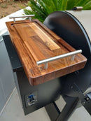 The Baby BBQ Board® 18" x 8" Cutting Board by BBQ Boards® features metal handles, a carved logo, and doubles as a stylish serving tray—shown resting on a black grill.