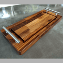 A wooden serving tray with metal handles holds the BBQ Boards® Baby BBQ Board® 18" x 8" Cutting Board. Both showcase varied wood tones and smooth finishes, elegantly arranged on a reflective gray surface.