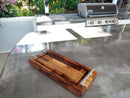 The BBQ Boards® Baby BBQ Board® 18" x 8" Cutting Board, featuring sleek metal handles, doubles as a stylish serving tray on your outdoor kitchen counter—ideal for your next Baby BBQ Board gathering with grill and greenery in the background.