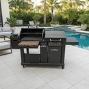 A BBQ Boards® Blackstone Pellet Grill Combo, featuring a front or side cutting board, stands on a tiled patio near a pool with palm trees and modern outdoor seating in the backyard.