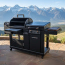 A BBQ Boards® Blackstone Pellet Grill Combo with Front or Side Cutting Board sits on a stone patio against scenic mountains and a blue sky, featuring clean surfaces and compartments—ready for your next outdoor feast.