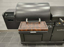 The BBQ Boards® Blackstone Pellet Grill Combo Front or Side Cutting Board by BBQ Boards® is shown on a metal tray at the front of a closed-lid pellet grill with digital controls, ideal for any prep station. The grill stands on a tiled floor against a light wall.