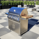 A stainless steel outdoor gas grill with four burners and a lid thermometer features a BBQ Boards® Blaze Grills LTE or LTE+ Side Cutting Board, set on a checkered patio with modern outdoor furniture and tall plants in the background.