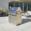 A BBQ Boards® Blaze Grills LTE Side Cutting Board is featured on a stainless steel outdoor gas grill, set on a modern patio with potted plants and outdoor seating in the background.