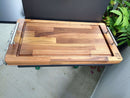 The BBQ Boards® GMG Daniel Boone/Ledge & Jim Bowie/Peak Side Cutting Board by BBQ Boards® sits on a countertop, showcasing metal handles, an edge groove, and a logo. Ideal for BBQ prep with plants in the background.
