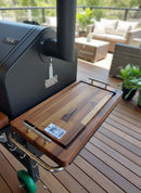 A BBQ Boards® GMG Daniel Boone/Ledge & Jim Bowie/Peak Side Cutting Board by BBQ Boards® is attached to a grill on a wooden deck with plants and patio furniture. A BBQ competition sticker decorates this convenient prep space.