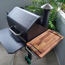 A black outdoor smoker grill with a chimney sits on a patio, featuring the BBQ Boards® GMG Daniel Boone/Ledge & Jim Bowie/Peak Side Cutting Board from BBQ Boards® attached for BBQ prep. Lush green plants create a vibrant backdrop.