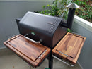 A black GMG Daniel Boone Ledge pellet grill with BBQ Boards® GMG Daniel Boone/Ledge front and side cutting boards is set up on a patio near green plants and a white railing.