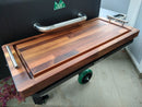 A BBQ Boards® GMG Daniel Boone/Ledge Front Cutting Board with metal handles sits on a grill shelf above concrete, enhancing your pellet grill workspace. Delicate pink flower petals to the left add a charming touch.
