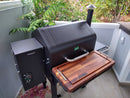 A black pellet grill with a BBQ Boards® GMG Daniel Boone/Ledge Front Cutting Board is on a patio by green plants and a gray fence. The grill has a digital control panel, chimney, and large workspace. Pink petals are scattered on the concrete floor.