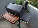 A black outdoor pellet grill with a side chimney and a BBQ Boards® GMG Daniel Boone/Ledge Front Cutting Board by BBQ Boards® for extra workspace, set on a concrete patio near a white fence and green plants.