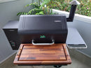 A black pellet grill features a BBQ Boards® GMG Daniel Boone/Ledge Front Cutting Board as a spacious workspace, with the control panel on the left and hooks on the right, set on a patio surrounded by greenery.