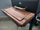 A BBQ Boards® GMG Daniel Boone/Ledge Front Cutting Board with metal handles sits on the side shelf of a black pellet grill, showcasing the BBQ Boards® logo and providing a convenient workspace in a covered patio area.