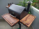 A covered patio with a black smoker grill features BBQ Boards® GMG Daniel Boone/Ledge Trio Front, Side & Pellet Bin Cutting Boards (set of three) by BBQ Boards®, Green Mountain Grills accessories, spice jars, and plants against a light gray wall.