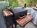 A black GMG Daniel Boone Ledge Trio grill features BBQ Boards® Front, Side & Pellet Bin Cutting Boards (Set of Three) on a patio with greenery, transforming the left shelf into a convenient prep station for your next BBQ.